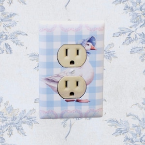 May include: A decorative light switch cover featuring a goose illustration. The cover has a blue and white gingham pattern with a pink lace trim. The goose is wearing a blue bonnet. The cover has two electrical outlets.