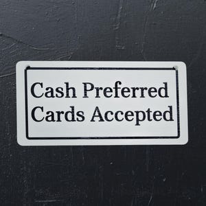 May include: A rectangular white sign with black text that reads "Cash Preferred Cards Accepted." The sign has a black border and is mounted on a dark gray textured surface.