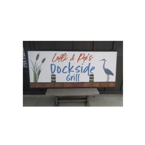 May include: A rectangular sign for "Lolli & Pop's Dockside Grill" with a white background, brown border, and brick-like bottom. The sign features a blue heron, cattails, and text in red and blue.