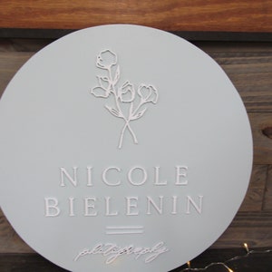 May include: A round, light blue sign with the name "NICOLE BIELENIN" in embossed lettering. The sign features a floral design and the word "photography" in script. The sign is likely for a photography business.