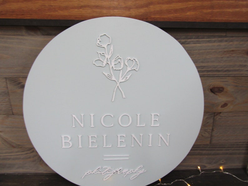 May include: A round, light blue sign with the name "NICOLE BIELENIN" in embossed lettering. The sign features a floral design and the word "photography" in script. The sign is likely for a photography business.