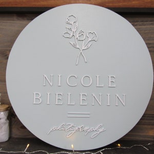 May include: A round, light blue wooden sign with the name "NICOLE BIELENIN" in raised white lettering. Below the name is the word "photography" in cursive. A floral design is at the top. The sign is a decorative item.