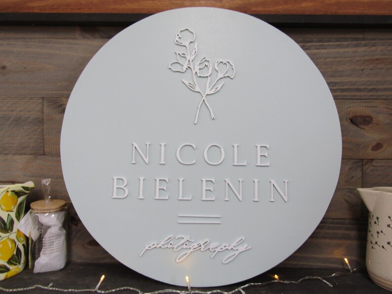 May include: A round, light blue wooden sign with the name "NICOLE BIELENIN" in raised white lettering. Below the name is the word "photography" in cursive. A floral design is at the top. The sign is a decorative item.