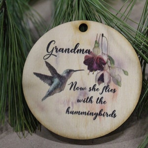 May include: A round wooden ornament with a hummingbird and flowers printed on it. The ornament has the text "Grandma" and "Now she flies with the hummingbirds".