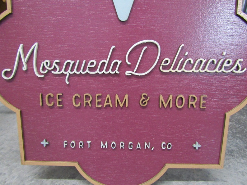 May include: A red wooden sign with a white cursive font that reads "Mosqueda Delicacies Ice Cream & More" and a white font that reads "Fort Morgan, CO".