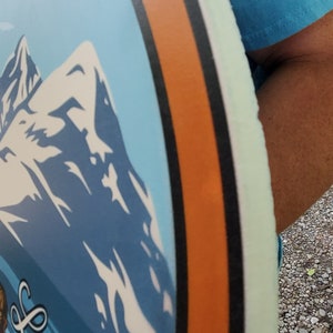 May include: A round, blue and white disc with a mountain scene and the word "Logan" written in white cursive. The disc has a black and orange border.
