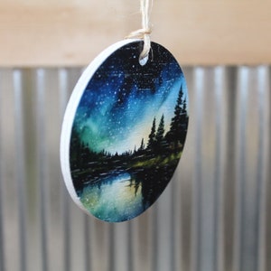 May include: Round wooden ornament with a painted scene of a forest at night with a starry sky and a reflection of the trees in a lake. The ornament is painted in shades of blue, green, and black.
