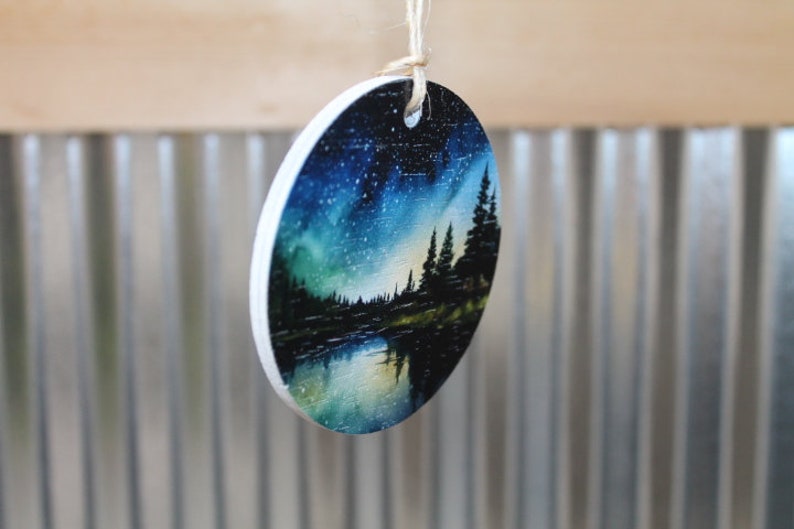 May include: Round wooden ornament with a painted scene of a forest at night with a starry sky and a reflection of the trees in a lake. The ornament is painted in shades of blue, green, and black.