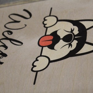 May include: A wooden sign with the word "Welcome" written in cursive script. A cartoon dog with black eyes and a red tongue is peeking over the top of the sign.
