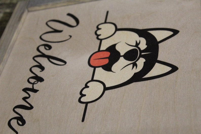 May include: A wooden sign with the word "Welcome" written in cursive script. A cartoon dog with black eyes and a red tongue is peeking over the top of the sign.