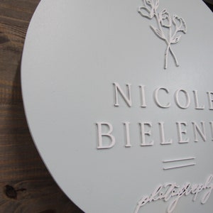 May include: A round, light blue sign with the name "Nicole Bielenin" in white, raised lettering. The sign also features a floral design and the word "photography" in script. The sign is mounted on a wooden background.