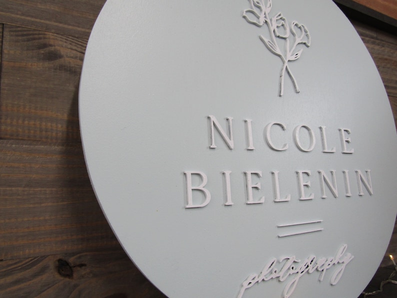 May include: A round, light blue sign with the name "Nicole Bielenin" in white, raised lettering. The sign also features a floral design and the word "photography" in script. The sign is mounted on a wooden background.