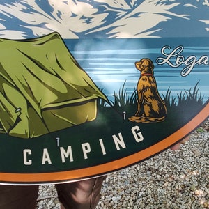 May include: A green tent with a mountain and lake scene in the background. The text "FAMILY CAMPING" is on the bottom of the sign. The name "Logan" is written in cursive on the top right of the sign. A brown dog is sitting in the grass on the right side of the sign.