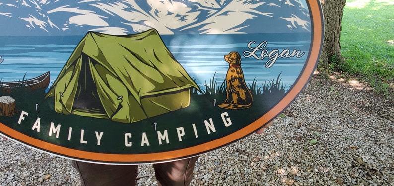 May include: A green tent with a mountain and lake scene in the background. The text "FAMILY CAMPING" is on the bottom of the sign. The name "Logan" is written in cursive on the top right of the sign. A brown dog is sitting in the grass on the right side of the sign.
