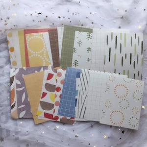 May include: A set of 12 decorative paper cards with various patterns. The cards feature geometric shapes, lines, dots, and nature-inspired designs in a muted color palette of yellow, orange, red, green, blue, and brown.