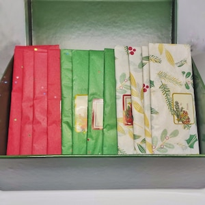 以下が含まれることがあります： 蓋が開いた緑色の箱の中に、和紙テープが12ロール入っています。和紙テープは、ホーリー、ベリー、雪片など、さまざまなクリスマスのテーマで飾られています。箱には「12 Days Washi Calendar」とラベルが貼られています。