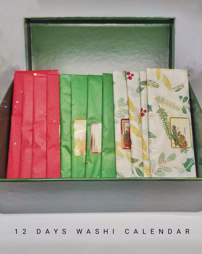 以下が含まれることがあります： 蓋が開いた緑色の箱の中に、和紙テープが12ロール入っています。和紙テープは、ホーリー、ベリー、雪片など、さまざまなクリスマスのテーマで飾られています。箱には「12 Days Washi Calendar」とラベルが貼られています。