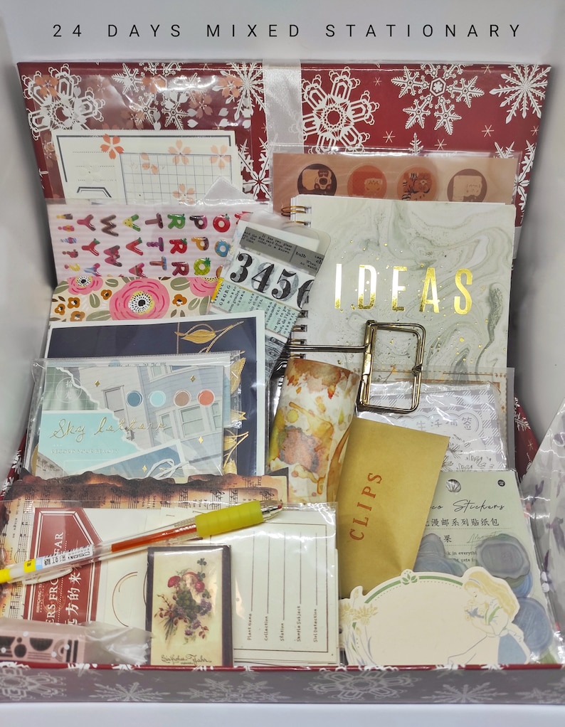 以下が含まれることがあります： 雪の結晶と「24 Days Mixed Stationery」の文字で飾られた赤と白の箱。箱の中には、ステッカー、紙、封筒など、さまざまな文房具が入っています。