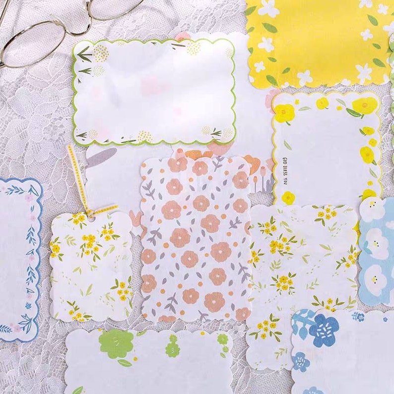 May include: Assortment of decorative paper note cards with scalloped edges and floral patterns. The cards are white with various floral designs in pink, yellow, blue, and green.
