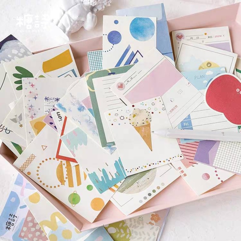 May include: A collection of colorful and patterned decorative paper stickers. The stickers feature various designs, including geometric shapes, ice cream cones, and abstract patterns. The stickers are arranged in a pink box.