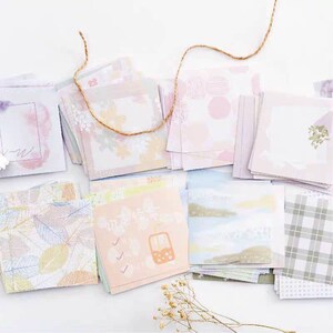 May include: A set of pastel colored decorative paper squares with various patterns, including leaves, flowers, and geometric shapes. The paper squares are arranged in a random pattern on a white surface.