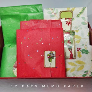以下が含まれることがあります： 前面に「12 Days Memo Paper」と書かれた赤い箱。箱の中には赤と緑の薄紙とメモ用紙の束が入っています。