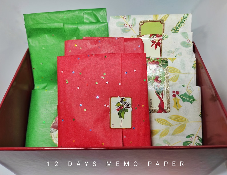 以下が含まれることがあります： 前面に「12 Days Memo Paper」と書かれた赤い箱。箱の中には赤と緑の薄紙とメモ用紙の束が入っています。