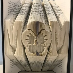 May include: A book art sculpture of the word "MOM" with a butterfly in the center. The book is open and the pages are folded to create the letters and the butterfly. The book is bound in black with a navy blue ribbon.