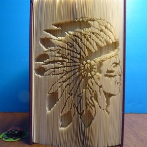 May include: A book with a 3D pop-up design of a Native American wearing a headdress. The book is closed and standing upright on a wooden surface.