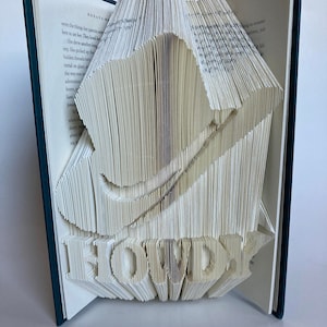 May include: A book with pages folded into a three-dimensional design of a cowboy hat with the word "HOWDY" underneath. The book is open and the pages are white.
