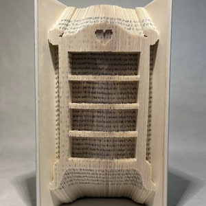 May include: A white book with a 3D book folding sculpture of a bookshelf. The bookshelf has five shelves and a heart-shaped cutout on the top.