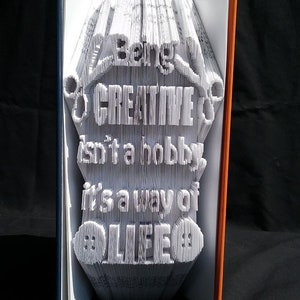 May include: A white book with the words "Being Creative isn't a hobby it's a way of life" folded into a 3D design. The book is open and the spine is blue and orange.