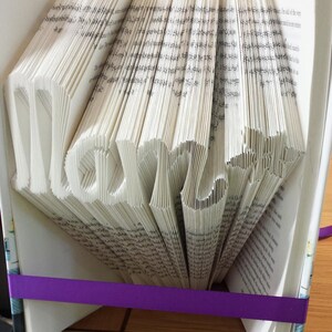 May include: A book with the word "Mom" folded into the pages. The book is bound in white with a purple ribbon around it. The book is sitting on a wooden table.