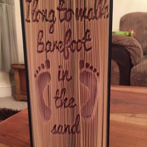 May include: A book with the words "Long to walk barefoot in the sand" folded into the pages. The words are cut out of the pages and the book is standing upright.