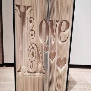 May include: A book with the word "Love" folded into the pages. The book is bound in black and has a white cover. There are three hearts folded into the pages.