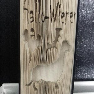 Könnte beinhalten: Ein Buch mit dem Text "Hallo-Wiener" auf dem Umschlag, gefaltet in Form von zwei Hunden. Das Buch steht aufrecht auf einer schwarzen Oberfläche.