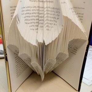 Puede incluir: Un libro doblado en forma de murciélago. El libro está abierto y las páginas están dobladas de forma que crean una forma tridimensional. El libro está sobre una superficie marrón claro.