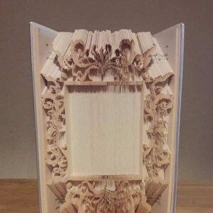 以下が含まれることがあります： 複雑なデザインで3Dフレームに折り畳まれた本。フレームは紙製で、薄い茶色です。フレームは木製の表面に立っています。