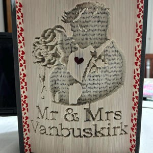 Peut inclure: Une sculpture d'art de livre représentant un couple qui s'embrasse, avec les mots "Mr. & Mrs. Vanbuskirk" sous l'image. La sculpture est réalisée à partir de pages d'un livre et est décorée de cœurs rouges.