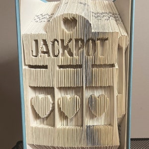May include: A book with the word "LUCKY" folded into the pages, with three hearts folded into the pages below. The book is open and standing upright on a black metal shelf.