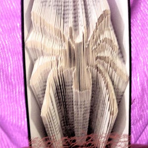 May include: A book folded into the shape of a spider, with the pages fanned out to create a three-dimensional effect. The spider is displayed on a pink fabric background with a brown net underneath.