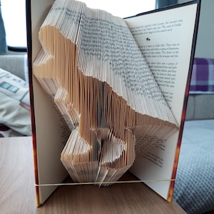 May include: A book with pages folded into the shape of a dinosaur. The book is open and the dinosaur is standing up. The book is held together with a rubber band.