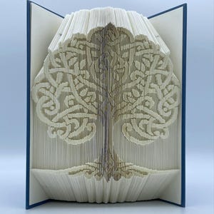 May include: A white paper book folded into the shape of a tree with intricate Celtic knotwork designs. The book is open and standing on a blue surface.