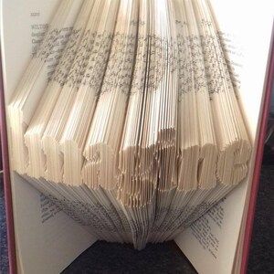 Könnte beinhalten: Ein Buch mit Seiten, die zu der Zahl "2023" gefaltet sind. Das Buch hat einen roten Einband und steht aufrecht auf einer dunklen Oberfläche.