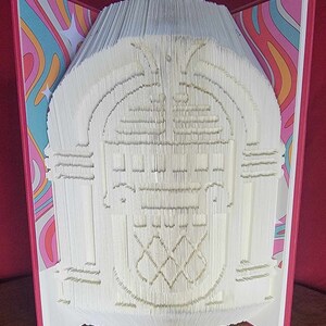 May include: A handmade book sculpture featuring a detailed jukebox design. The pages are intricately folded and cut to create the image. The book has a red cover and the inside pages have a colorful, retro pattern.