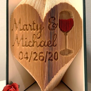 May include: A book folded into the shape of a heart with the words "Marry Michael" and the date "04/26/20" on the front. A red wine glass is folded into the heart. The book is standing on a white surface with a red ribbon and a red rose.