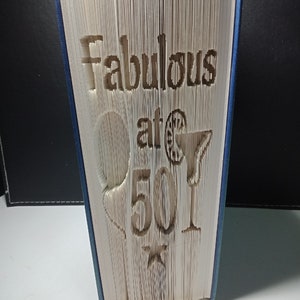 Op de afbeelding: Een wit boek met de woorden "Fabulous at 50!" uitgeknipt uit de pagina's. Het boek staat rechtop op een wit oppervlak.