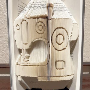 Op de afbeelding: Een beige boeksculptuur van een naaimachine, gemaakt door de pagina's te vouwen. De sculptuur toont gedetailleerde uitsparingen en vormen die de onderdelen van de machine voorstellen. Het boek is open en toont de tekst.