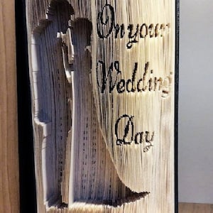 Könnte beinhalten: Ein Buch mit Seiten, die zu einer Silhouette eines Paares gefaltet sind, mit dem Text "On your Wedding Day" auf dem Einband.