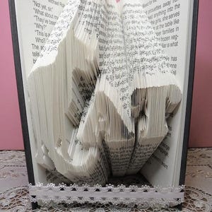 May include: A book art sculpture of the word "LOVE" created by folding the pages of a book. The book is open and the pages are folded to create the letters. The book is sitting on a white lace tablecloth with a ladybug figurine in front of it.